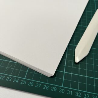 A stack of paper sheets rests on a green cutting mat. To the right is a bone folder.